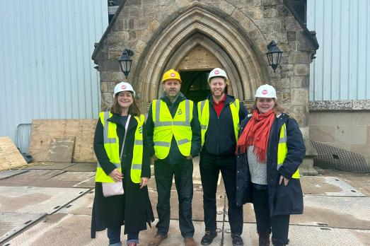 Four people in high-vis jackets and hard hats stand by a church