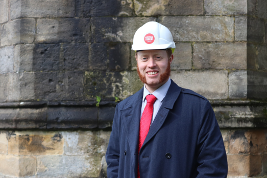 A headshot of a man with sideburns wearing a hardhat, red tie and navy coat.