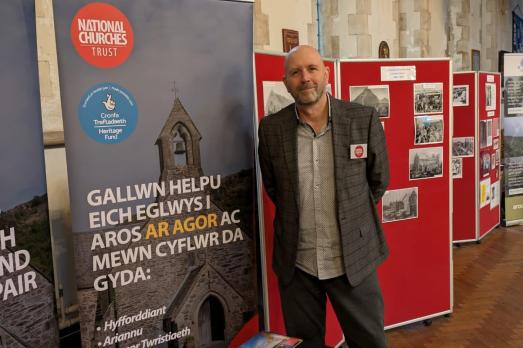 Gareth Simpson, our Welsh Support Officer, stands at a table filled with 'get support' leaflets and next to a roll up banner describing how the National Churches Trust helps churches.