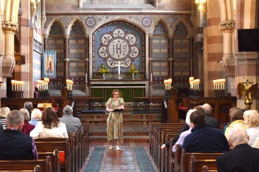A girl in a floral dress stands at the front of an ornate church aisle with people in pews facing her