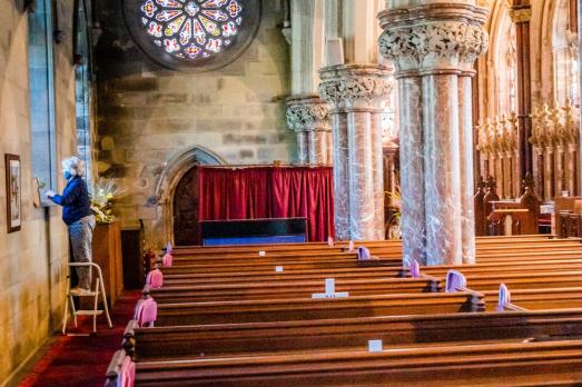 Rows of wooden pews with stone arched columns rising up on the right hand side. There's a stained glass window at the back of the photo and a woman stands on a ladder looking out. Interior of St Margaret Antioch