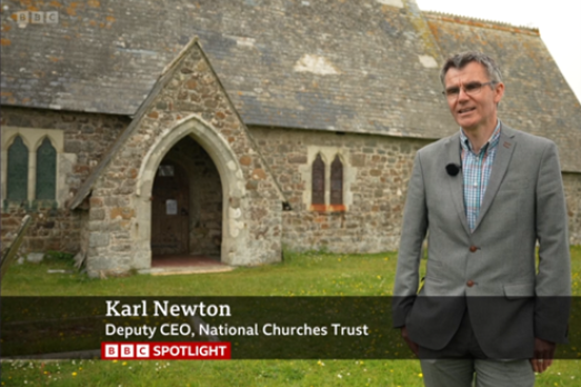 Karl Newton, Deputy CEO of the National Churches Trust, is wearing a grey suit and is standing outside of a very old church - St Grada & Holy Cross - in Cornwall. This is a screenshot from a BBC Spotlight interview.