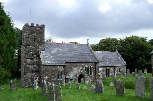Exterior of St Martin church. A historic church in Devon in grey stone with a square tower to the left of the image. Graves and green grass are in the foreground and a white and cloudy sky is behind the building.