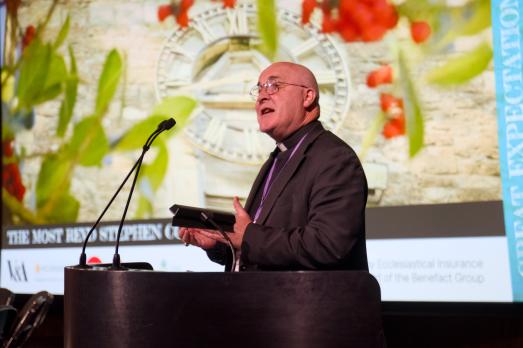 The Archbishop of York stands in front of a lectern delivering a speech. There is a colourful backdrop of a church and flowers behind him.