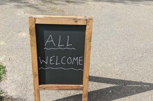 'All Welcome' written on a chalk board on the street (outside a church not in shot)