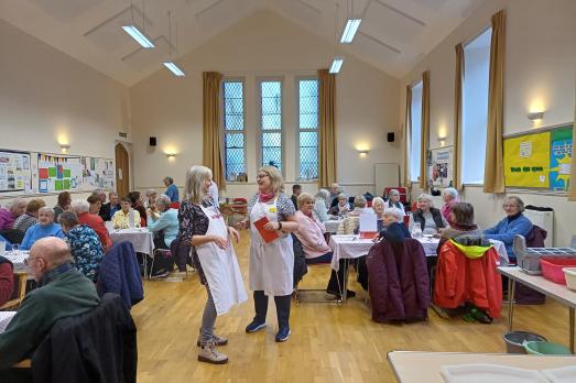 Two women in a church hall laugh. They are surrounded by many people eating while sat down at tables; a community hub.