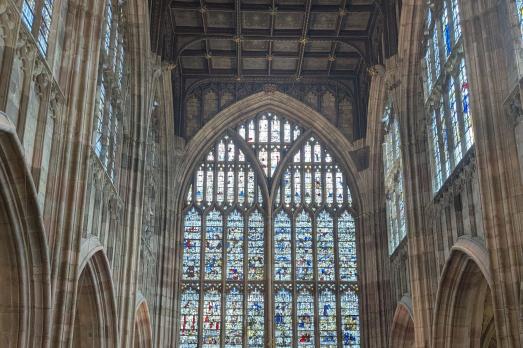 A photo of some of the beautiful stained glass at Great Malvern Priory. You can see one large arched window and several smaller windows on the left and right hand side.