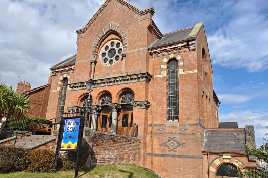 A large red bricked church on a hill with a blue sign out the front. Photo taken on a bright day.
