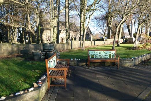 In the foreground are two benches; the stone church is the background of the shot with trees surrounding it