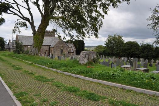 St Helen, a grey stone church, is partly obscured by a tree. There is a large graveyard in the foreground.