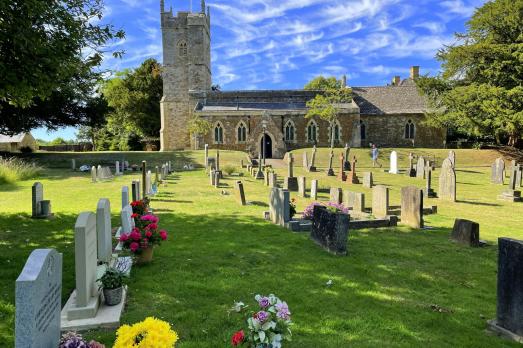 A beautiful stone church with a tower on the left-hand side and photographed on a bright and sunny day. There is a large graveyard in front of the church with flowers on many of the graves.