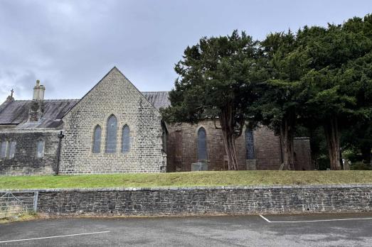 A grey stone Welsh church with many trees to the right-hand side of the image, which are party obscuring the church.