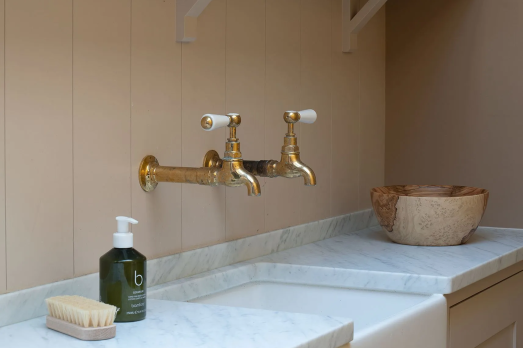 The image shows a marble sink with two gold taps above, soap next to it and a wooden bowl.