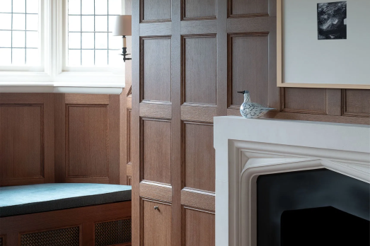 The image shows a bay window with a seat underneath, brown panelled walls and a black fireplace with a painting above.