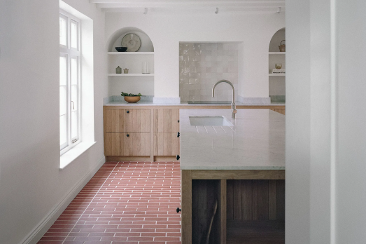 The image shows a kitchen with red floor tiles, a white marble island and wooden cabinets. There is a tall window letting light into the kitchen.