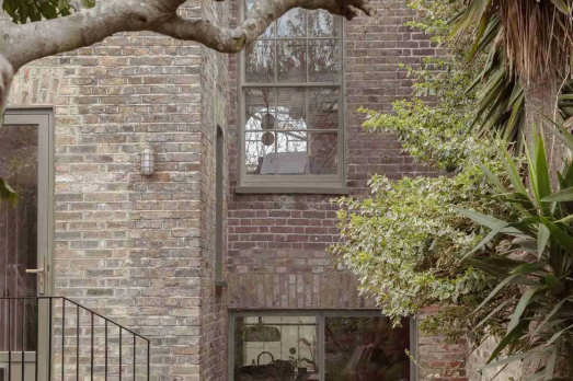 The image shows the outside of a house. The brick is grey and light brown. There are three windows and a staircase leading outside. There is green foliage and the branch of a tree over the picture.