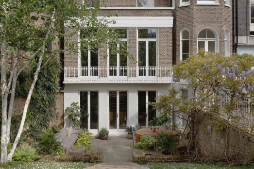 The image shows the outside wall of a house. It has tall windows and a garden table outside. There are two trees and some shrubs to the side of the house.