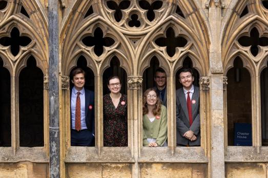 A group of five people stand in the cloisters of Lincoln Cathedrals - you can see them smiling in between intricate stone arches.