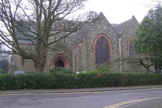 A stone church with arched windows. A row of bushes and trees are in the foreground.