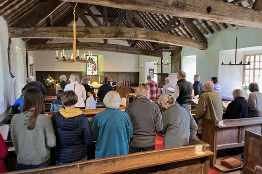 A group of people standing in pews facing the altar. A lovely beamed ceiling is visible.