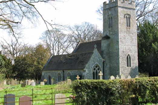 A grey stone church pictured from the pavement with a large stone tower to the front. The church is surrounded by greenery