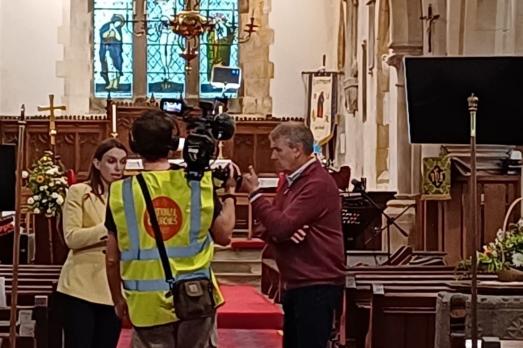 A man wearing a high vis vest films two people talking in a church
