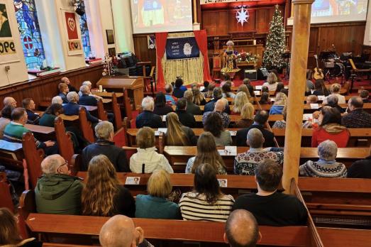 Rows of people in wooden pews face the front of the church. There is a Christmas Tree near the altar.