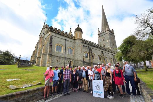 A group of people stand in front of a cathedral in Northern Ireland waving flags and standing in front of a The National Lottery Heritage Fund board