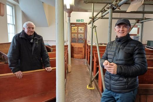Two men smile inside a Welsh chapel that is undergoing repairs to the ceiling