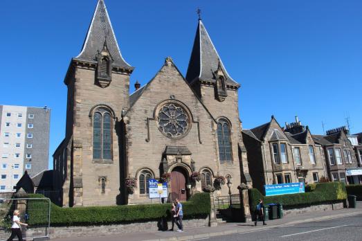 Taken from a distance, you can see a unique stone church with two large turrets either side of the entrance. People are walking on the path outside.