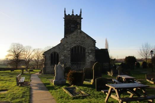 A picnic bench and graveyard photographed at dusk/dawn with St Peter's stone church and tower in the background