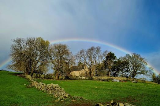 An arched rainbow over a grey-stone church partially obscured by trees