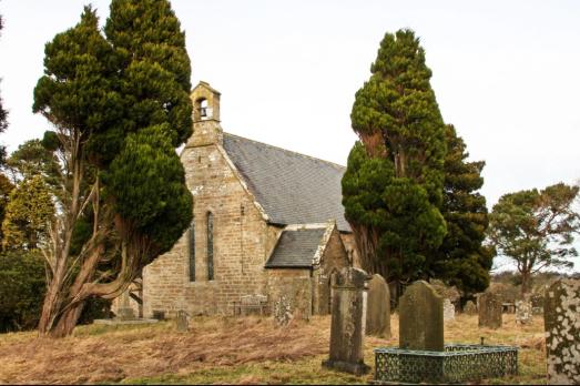 A stone church is visible between two trees. There re some graves in the foreground