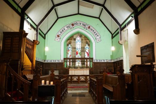 The interior of All Saints in Muggleswick; photograph is of the altar with a stained glass window in the middle. A wooden organ is to the side of the frame.