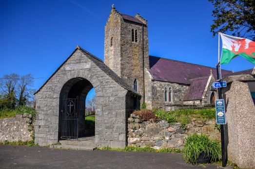 A stone church in the background; in the foreground is a Welsh flag waving in the wind and a stone pointed archway 