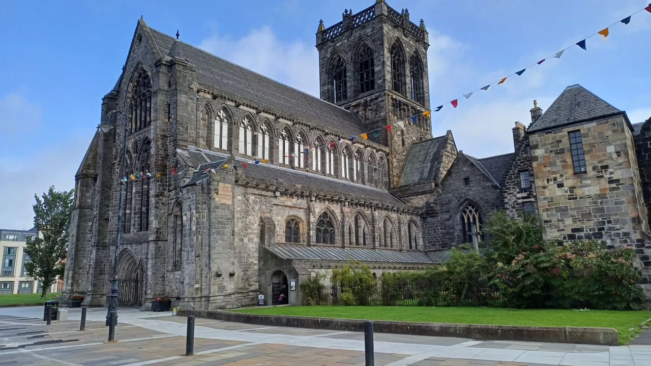 Image of a large grey stone abbey with a large central tower, blue skies, green grass and colourful bunting.
