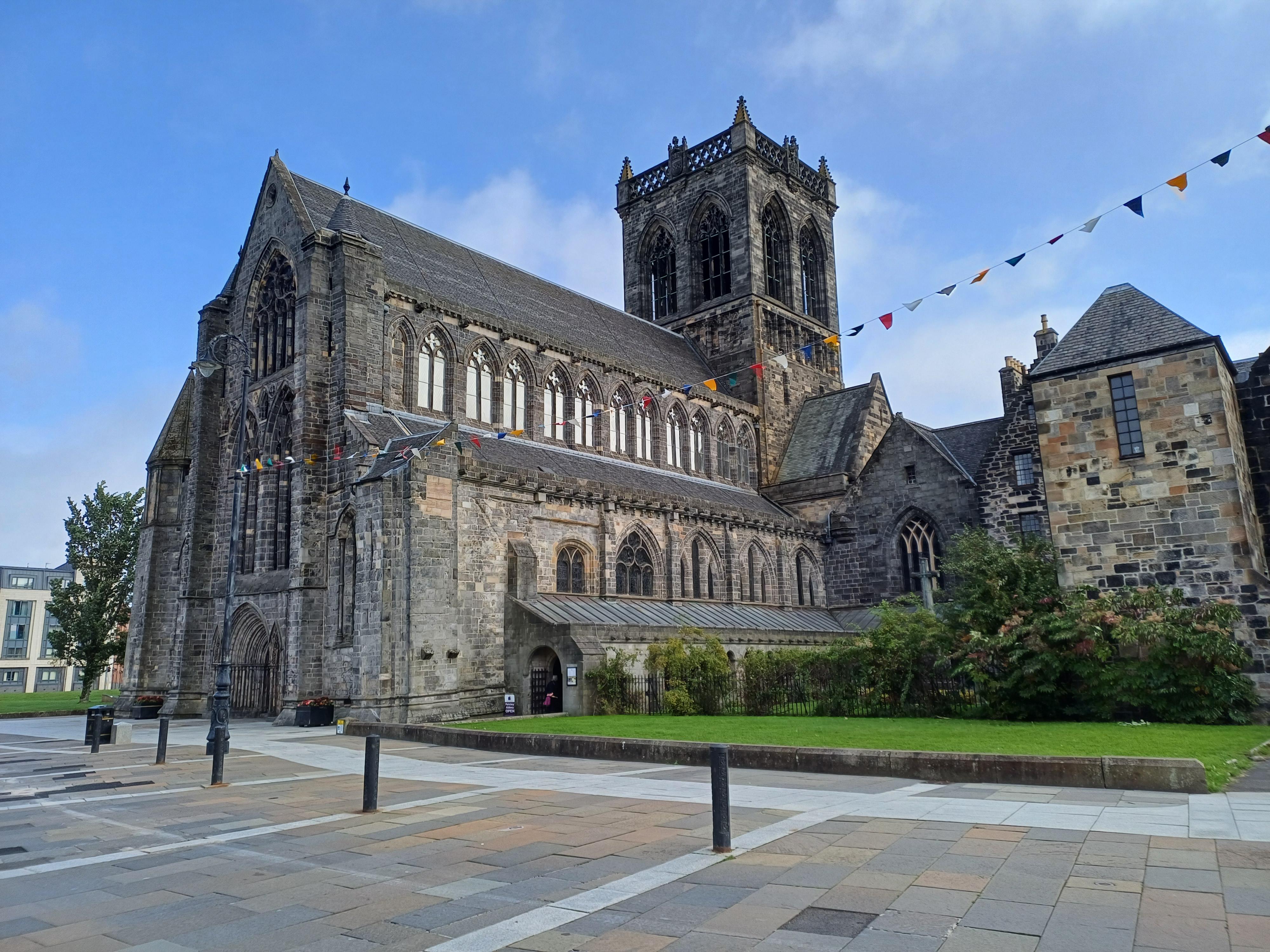 Image of a large grey stone abbey with a large central tower, blue skies, green grass and colourful bunting.
