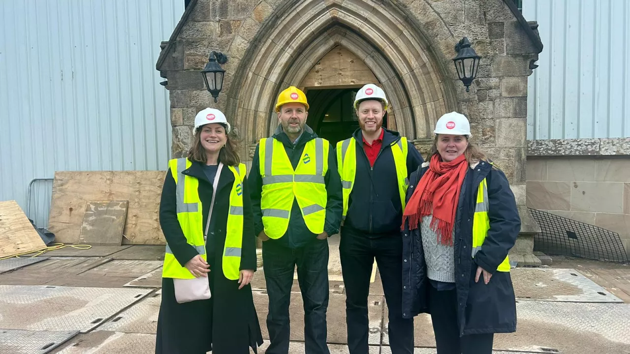 Four people in high-vis jackets and hard hats stand by a church