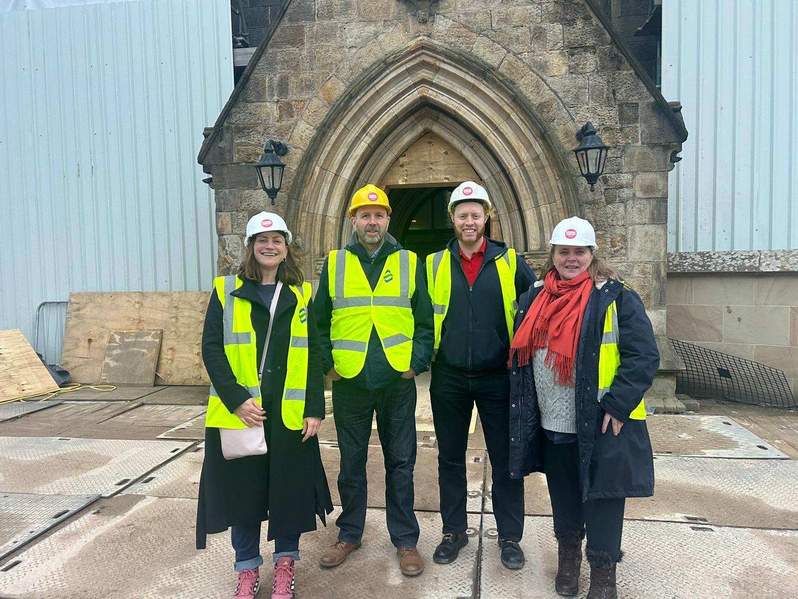 Four people in high-vis jackets and hard hats stand by a church