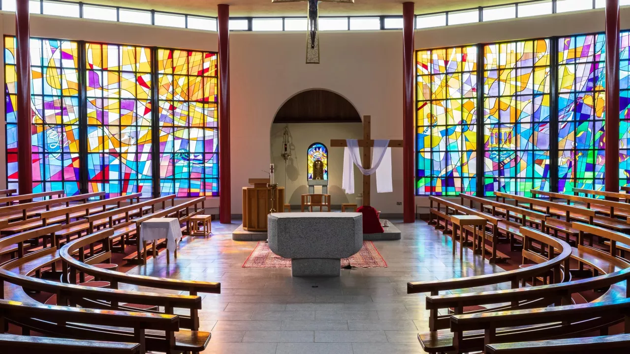 Interior of Carnlough St John the Baptist facing the altar with stained glass windows either side. Benches are in a circular fashion, facing the altar.