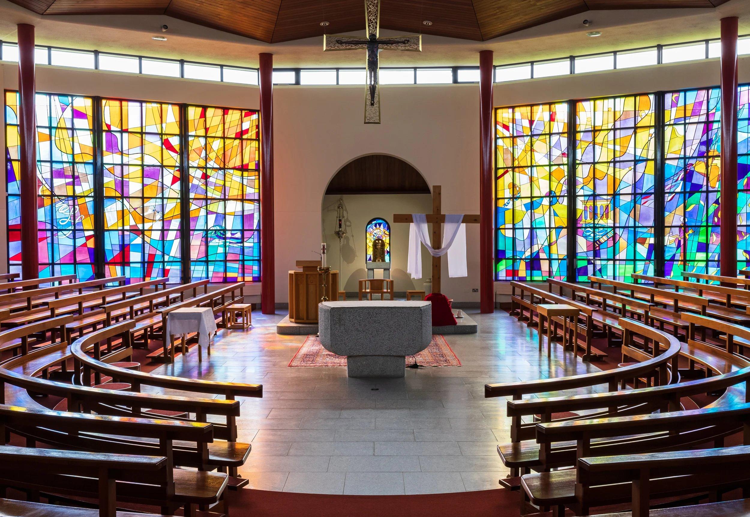 Interior of Carnlough St John the Baptist facing the altar with stained glass windows either side. Benches are in a circular fashion, facing the altar.