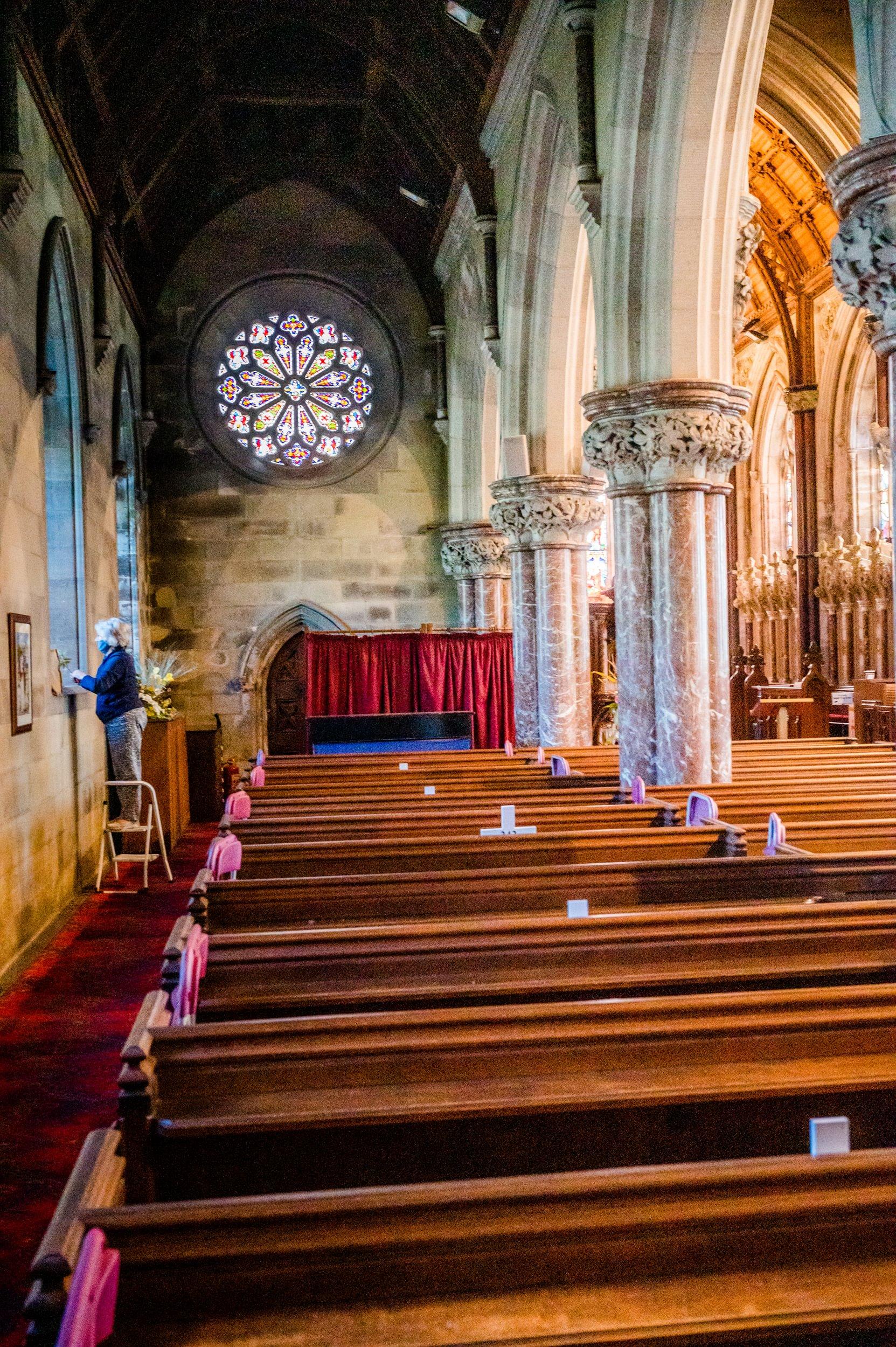 Rows of wooden pews with stone arched columns rising up on the right hand side. There's a stained glass window at the back of the photo and a woman stands on a ladder looking out. Interior of St Margaret Antioch
