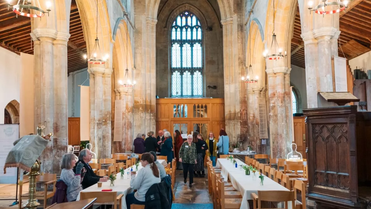 A pulpit to the right of the image and in the centre there are two tables in the aisle of the church with people sat at them as well as milling around.