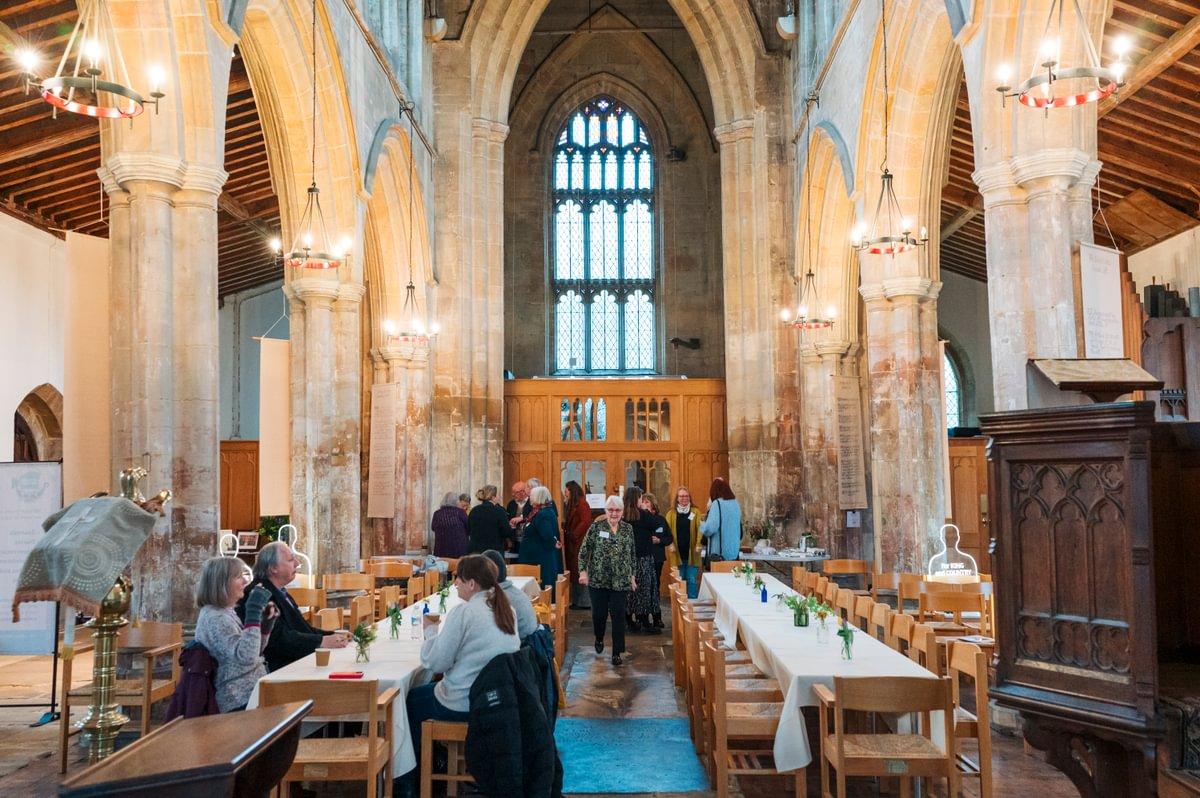 A pulpit to the right of the image and in the centre there are two tables in the aisle of the church with people sat at them as well as milling around.