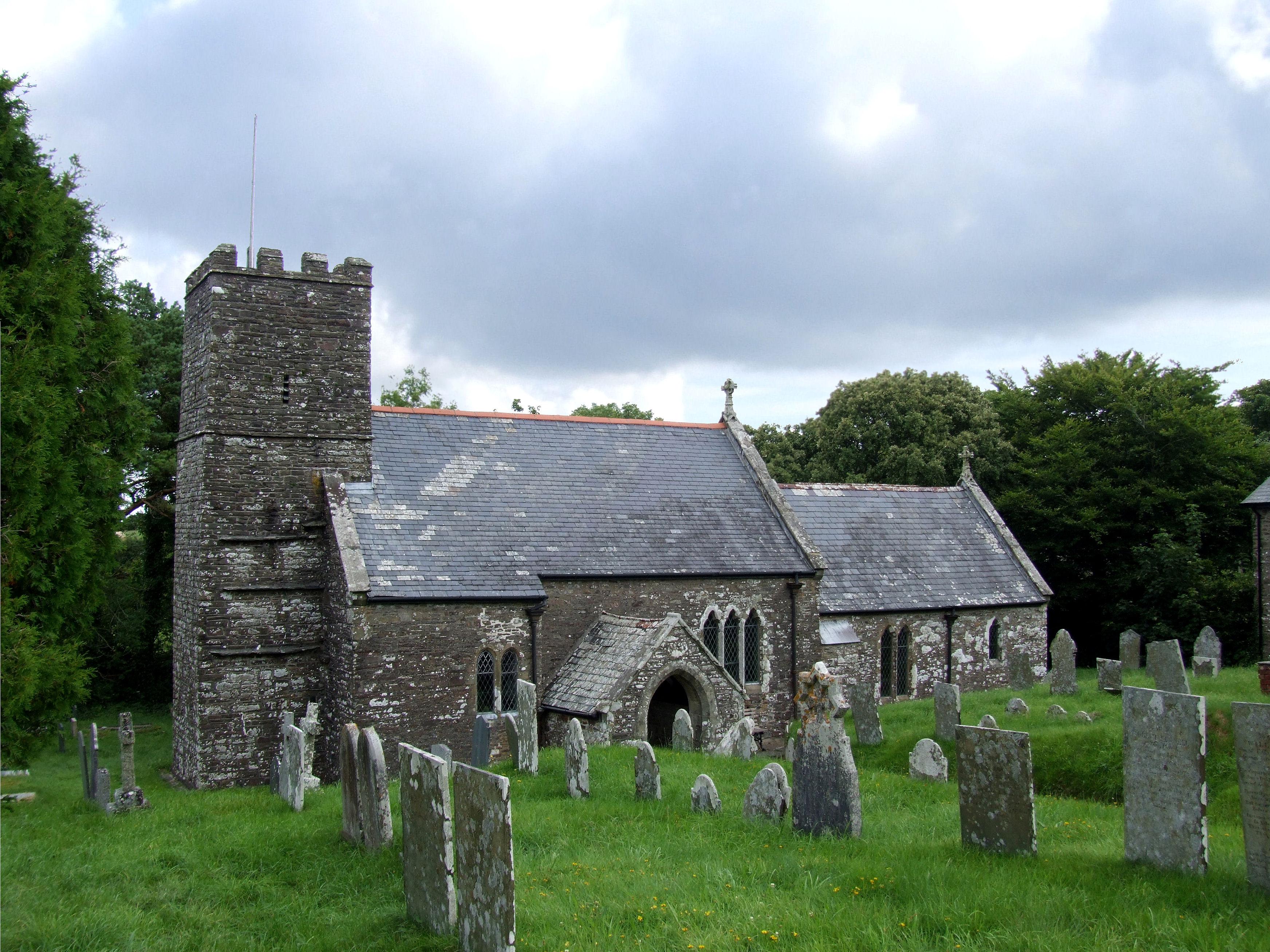Exterior of St Martin church. A historic church in Devon in grey stone with a square tower to the left of the image. Graves and green grass are in the foreground and a white and cloudy sky is behind the building. 