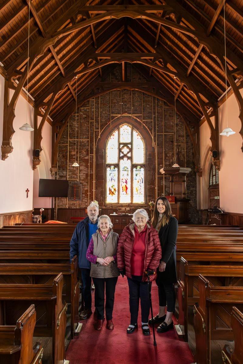 Four of the trustees of Tundergarth church inside the building. You can see stained glass in the background and pews on either side of the aisle.