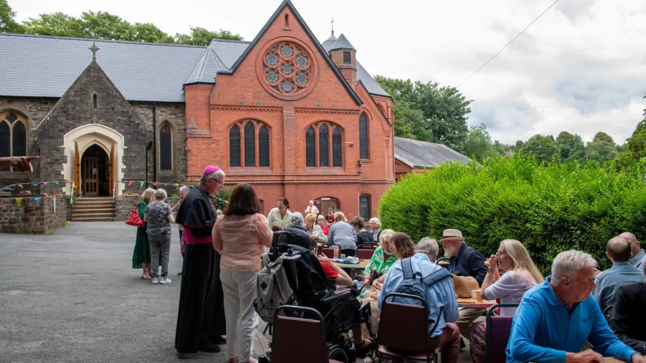 Table are set up outside the entrance to St Illtyd church to mark a celebration event. People are sitting down and milling around the tables. The church is in the background.