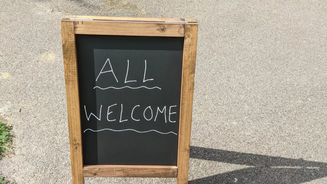 'All Welcome' written on a chalk board on the street (outside a church not in shot)