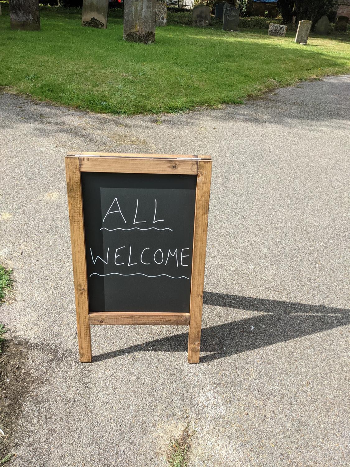 'All Welcome' written on a chalk board on the street (outside a church not in shot)