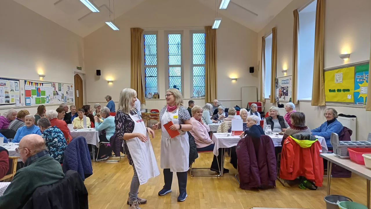Two women in a church hall laugh. They are surrounded by many people eating while sat down at tables; a community hub.